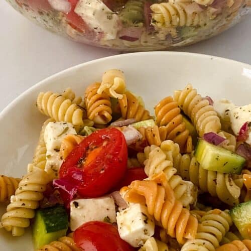 A bowl of rotini pasta salad featuring tri-color rotini tossed with cherry tomatoes, cucumber, red onion, and cubes of feta cheese. In the background, a larger serving bowl filled with the same vibrant rotini pasta salad completes the inviting scene.
