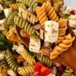 Tri-color Rotini Pasta Salad with feta cheese cubes, chopped red onion, cherry tomatoes, and cucumber served in a wooden spoon.