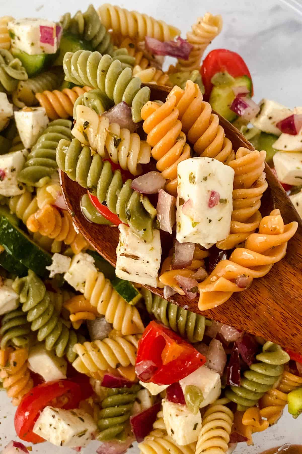 Tri-color Rotini Pasta Salad with feta cheese cubes, chopped red onion, cherry tomatoes, and cucumber served in a wooden spoon.