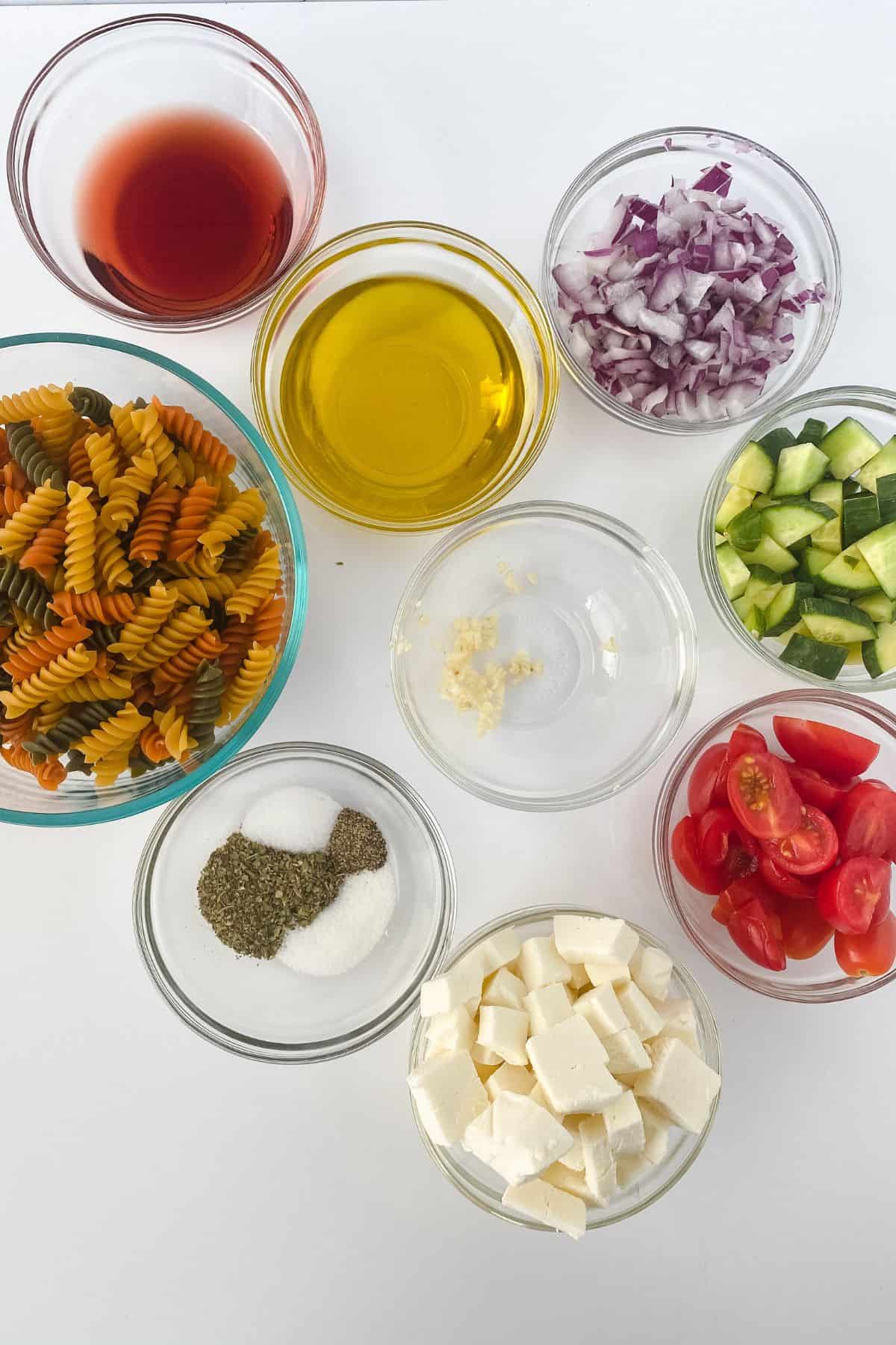 Nine bowls containing the fresh ingredients: red wine vinegar, olive oil, chopped red onions, tri-color rotini pasta, minced garlic, diced cucumber, cherry tomatoes, feta cheese cubes, salt, and black pepper.
