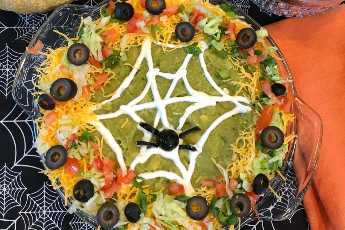On a black tablecloth with spider web designs on it, there is an orange piece of material on the right hand side and a glass dish with taco dip. The dip has a round border made of cheese, tomatoes, lettuce and black olives. In the middle there is guacamole that has a spider web drawn on it with sourcream and a spider made from a cut up black olive.  