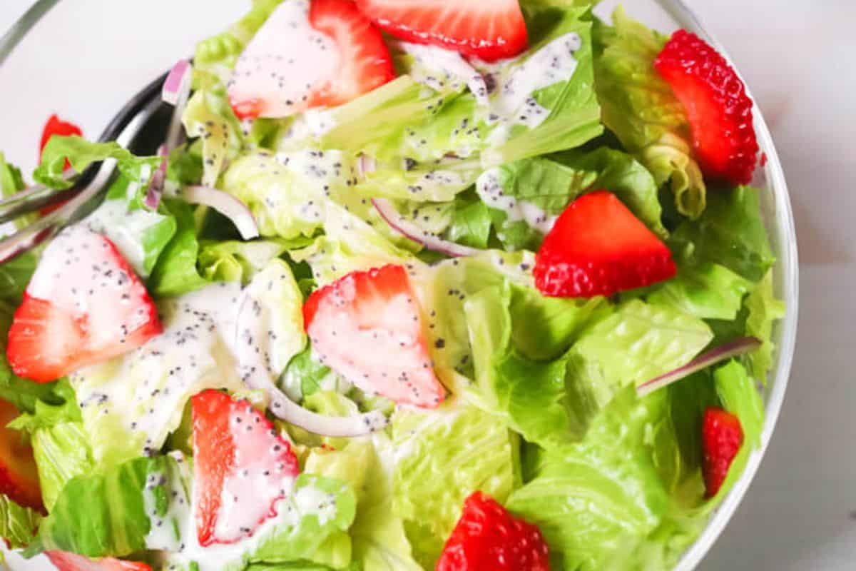 A close up of bright green lettuce topped with vibrant red strawberries and red onions cut into slices. The salad is topped with a white dressing with poppyseeds in it. 