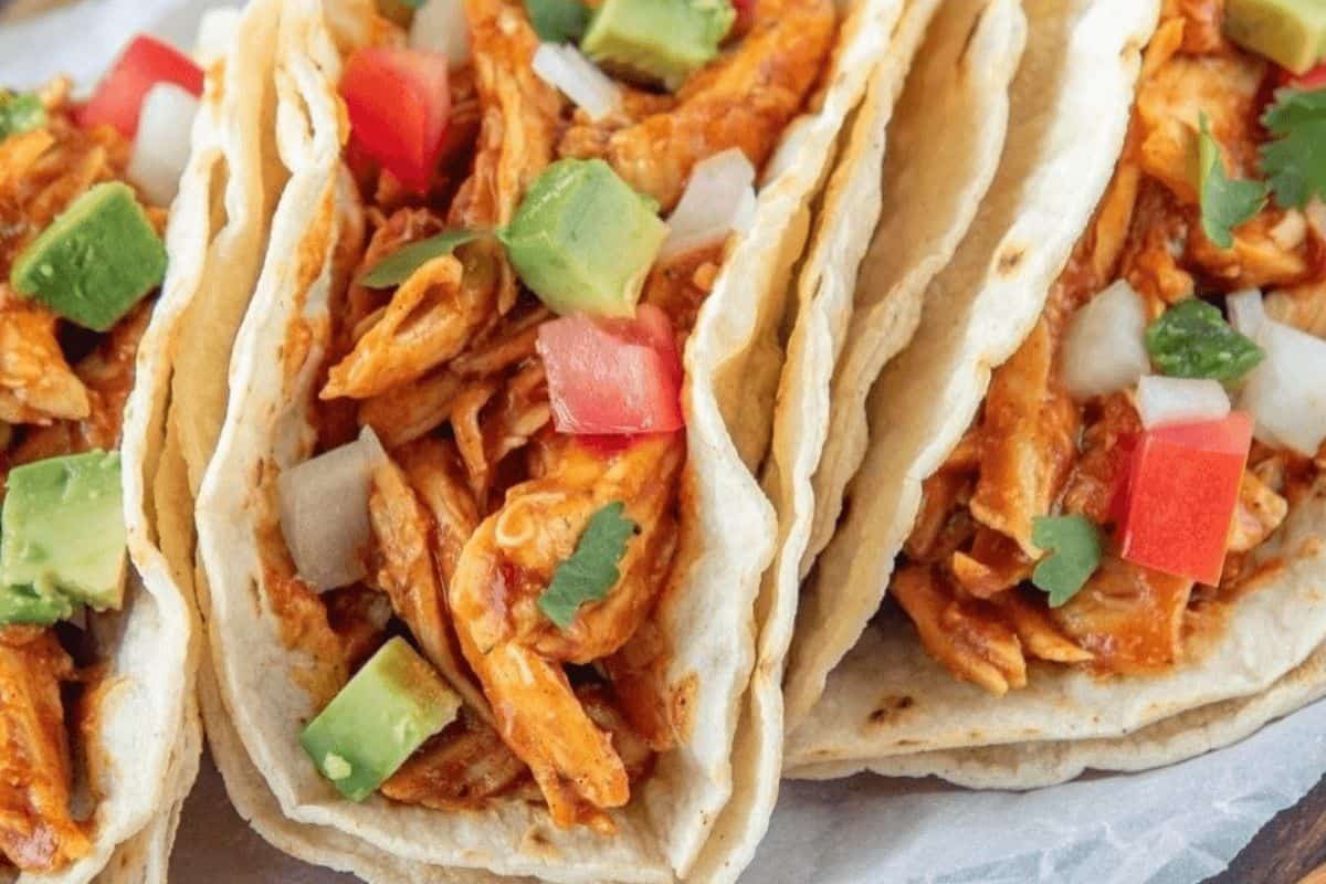A close up of soft tacos filled with strips of chicken in a red sauce and topped with diced tomatoes, onions, avocado and pieces of green seasoning. 
