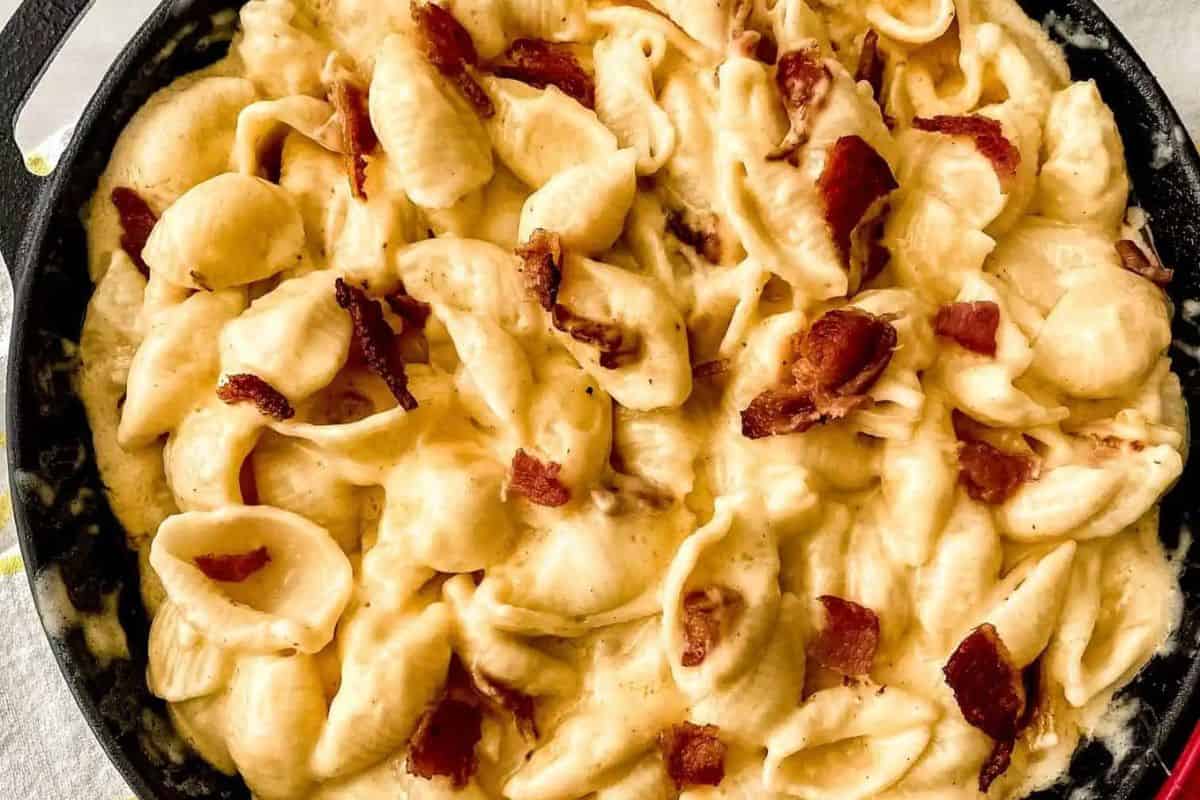 In a cast iron skillet, there is a close up of pasta shells covered with a creamy, yellow cheese sauce and bits of bacon. 