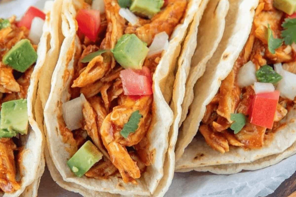 Three tacos tightly stacked together on a white wax paper background. The tacos are filled with shredded chicken in red sauce, diced tomatoes, diced onions, diced avocados and cilantro. 