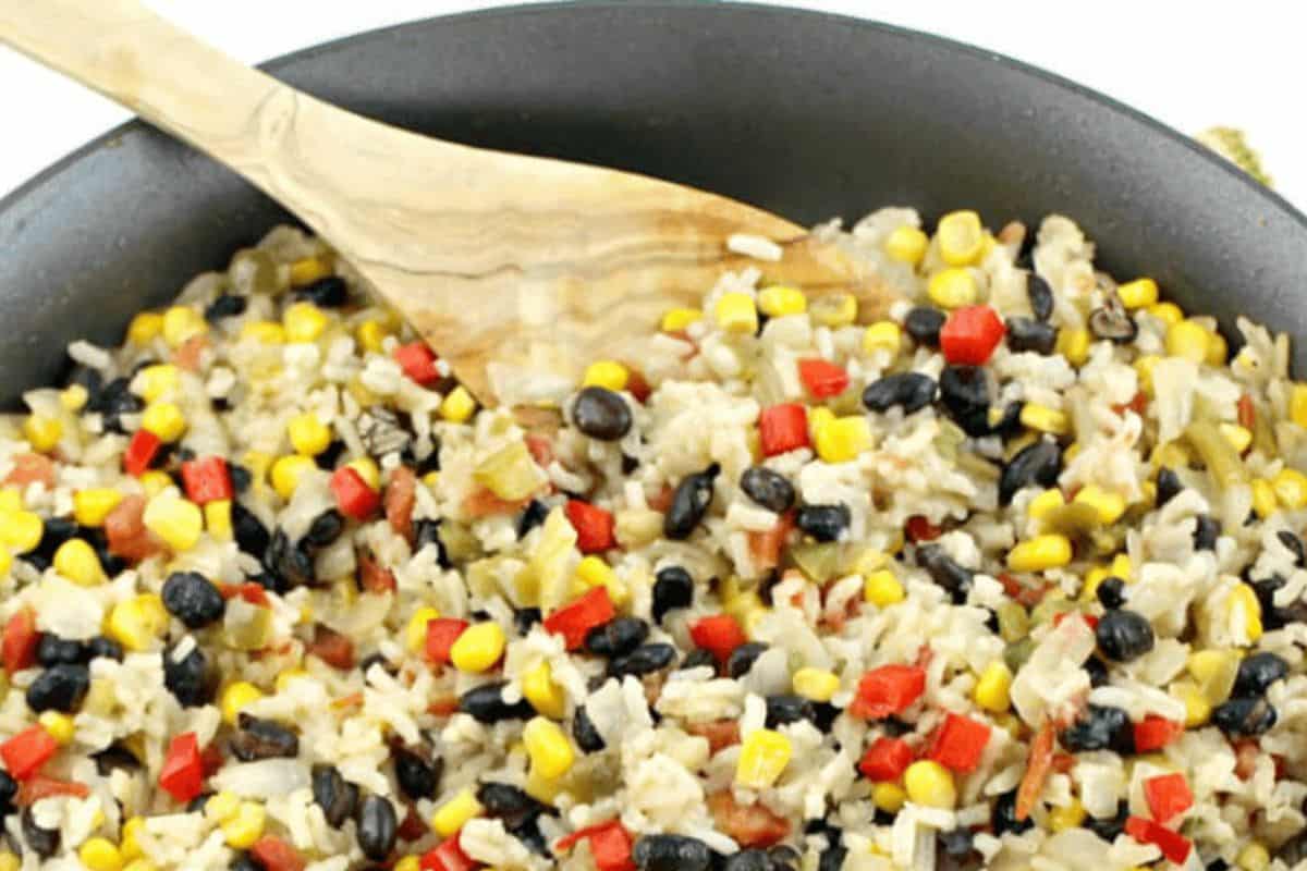 A black skillet, with a wooden spoon at the top of the picture, is filled with rice and beans. The dish is made up of off white rice, black beans, bright yellow corn, green chiles and vibrant red peppers. 