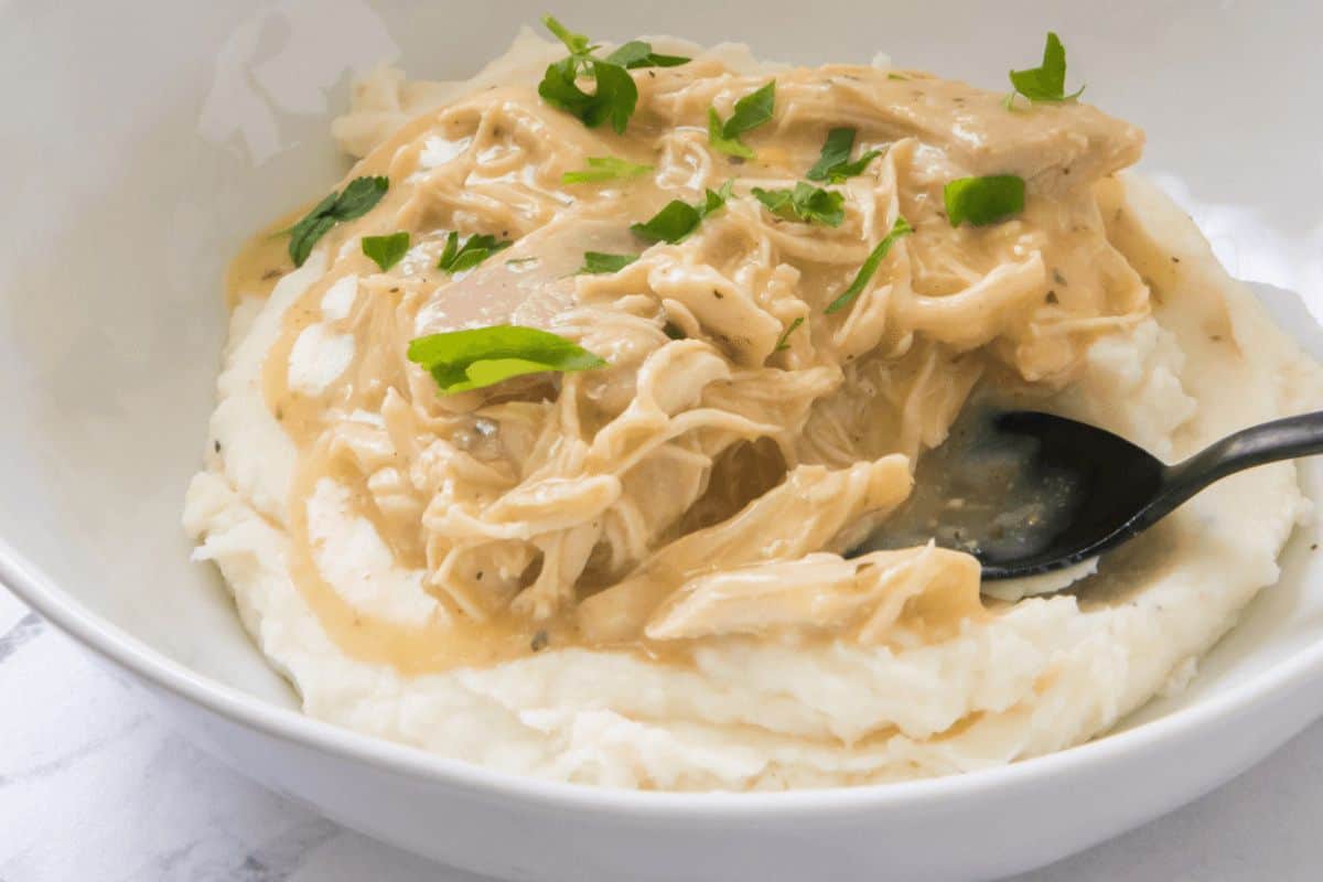 A close up picture of mashed potatoes topped with chicken and parsley. The chicken is shredded and in a gravy. A black spoon is stuck in the mashed potato on the right hand side of the picture. 