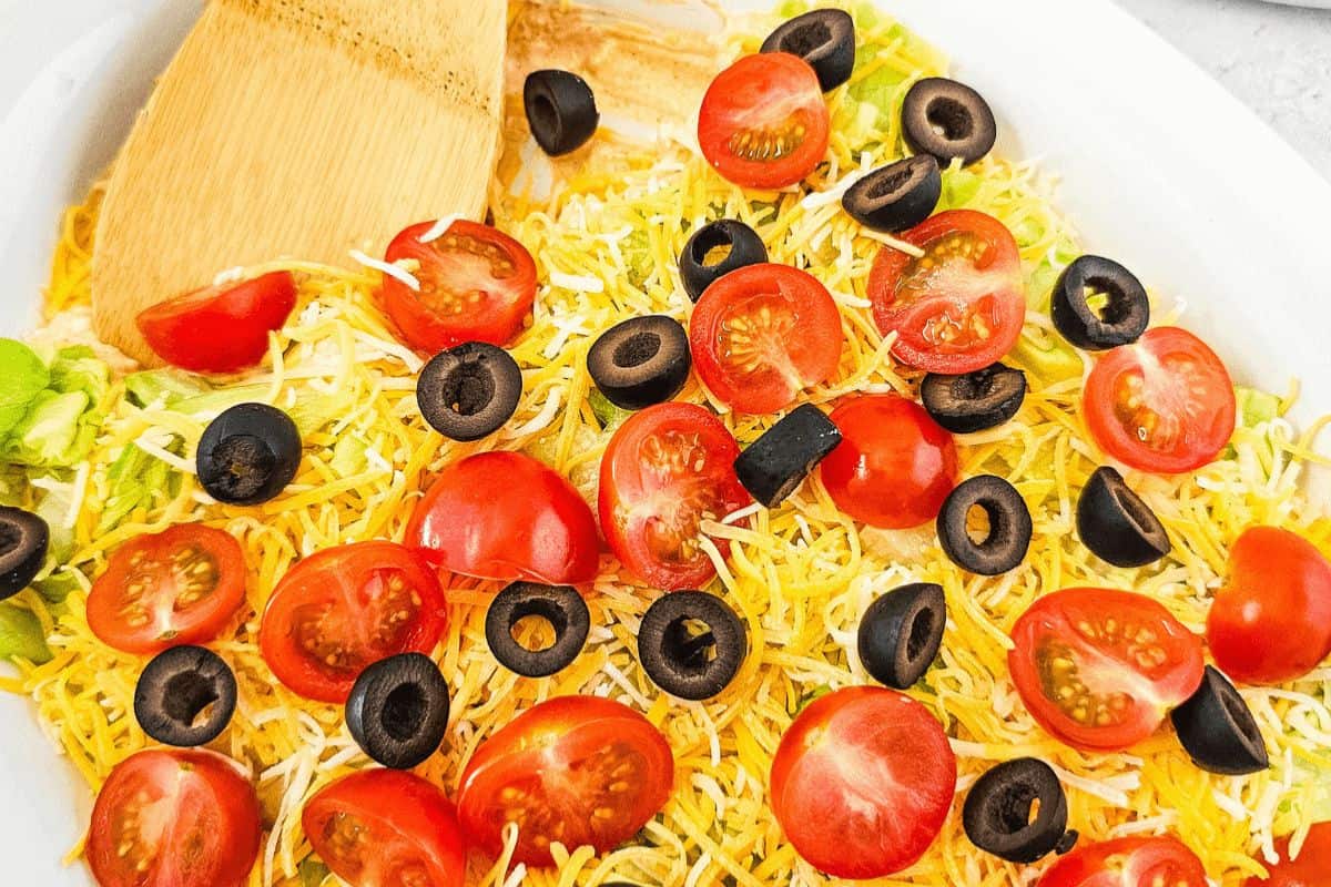A close up of a white baking dish filled with dip and topped with grated cheese, halved cherry tomatoes, halved black olives and shredded lettuce. In the top left of the picture there is a wooden spoon inserted in the dip. 