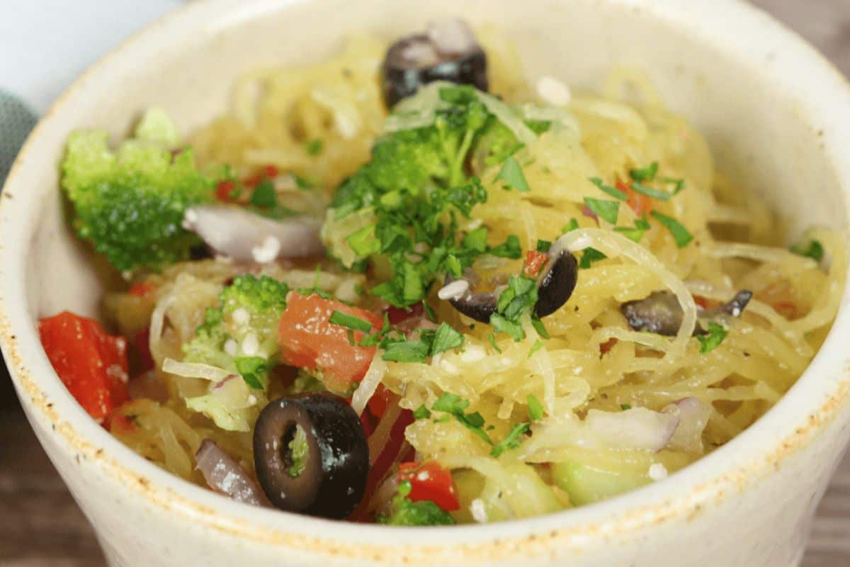 A very close up picture of spaghetti squash in an off white bowl. The spaghetti salad has spaghetti squash, tomatoes, broccoli, diced peppers, onions and black olives. It is topped with green herbs and sesame seeds. 