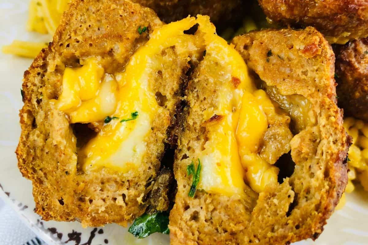 A very close up picture of meatballs on a white background. The meatball closest to the foreground of the picture has been halved so that you can see inside it. Melted cheese oozes out of the middle of the meatball. 