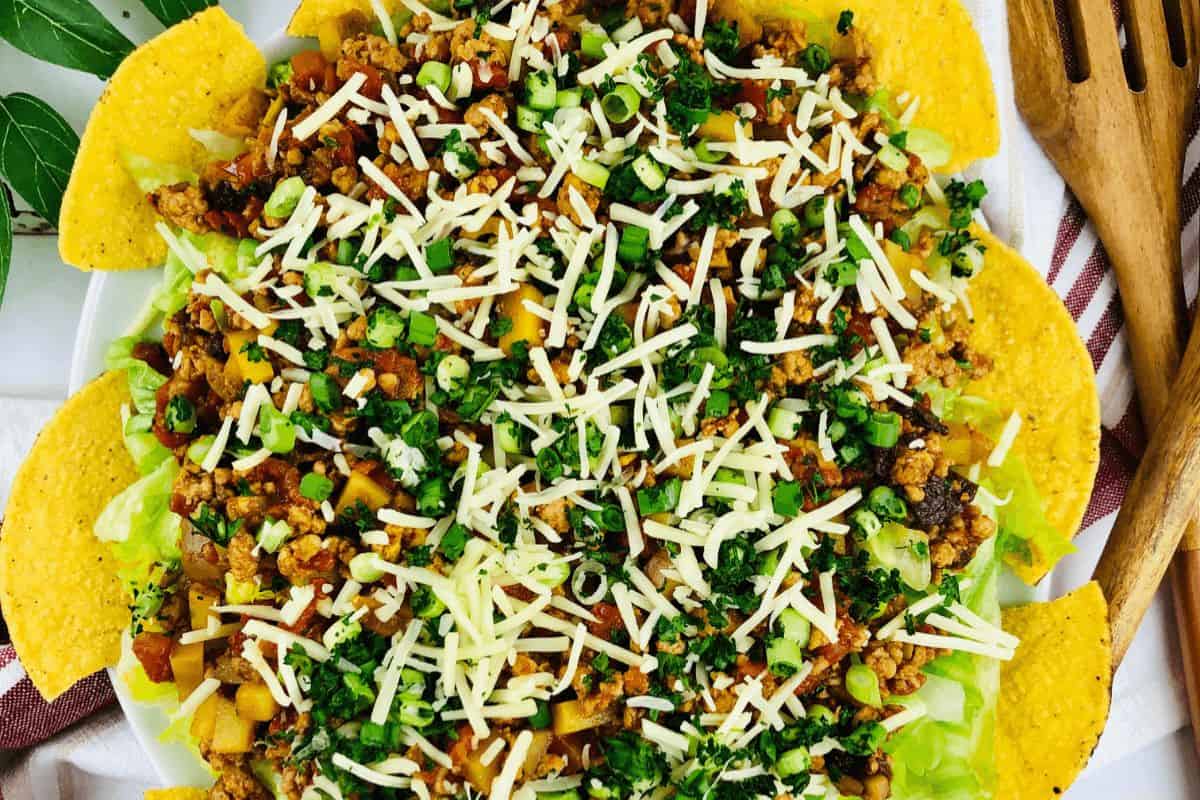 On a white plate, there are tortilla chips making a circle around the rim of the plate. On the inside there is a salad made of lettuce, ground chicken, green onions, diced olives, diced potatoes, diced tomatoes, cilantro and topped with grated cheese. To the right of the plate there is a set of wooden salad servers on a dark red and white striped tea towel.  