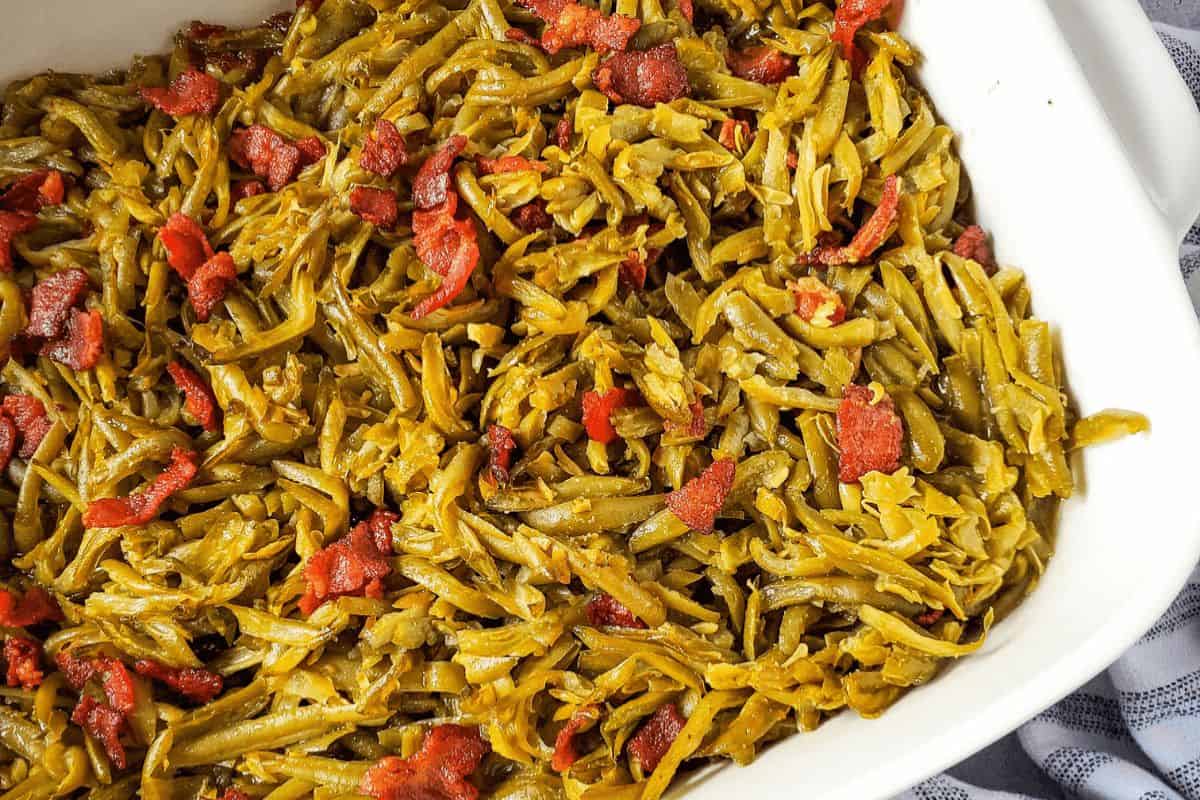 A close up of bacon green beans in a white casserole dish. The green beans look moist and are a deep green color. There are generous sprinklings of bacon across the top of the beans. 