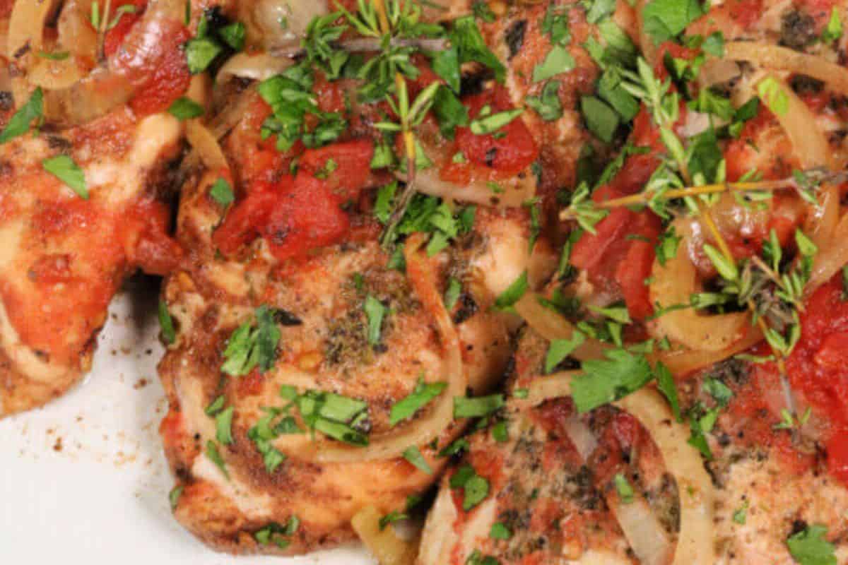 A close up of three chicken breasts covered with Italian seasonings, red sauce, tomatoes, onions, sprigs of thyme and chopped parsley. The chicken looks really moist and full of flavor. 