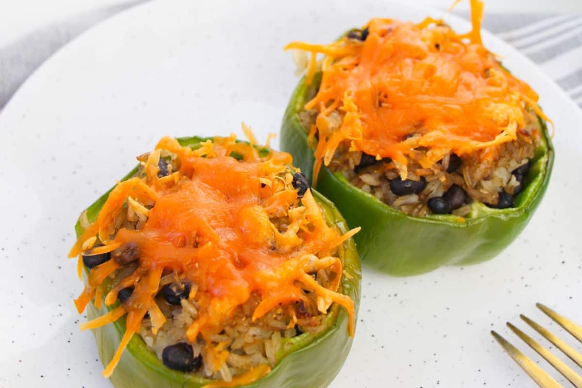 Placed in the middle of a white speckled plate are two green peppers. On the bottom right hand corner of the picture the tines of a gold fork are visible. The peppers have the tops cut off and are filled with rice, black beans and topped with melted cheese.