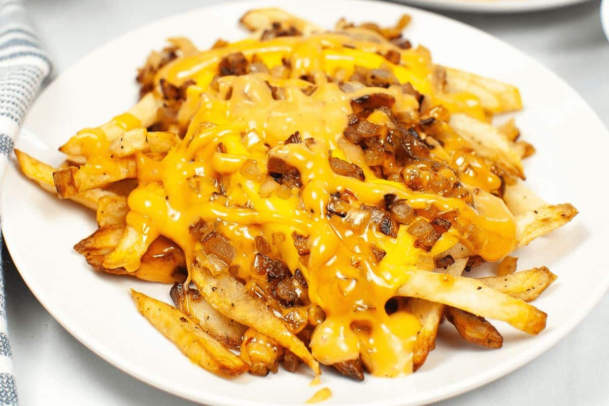 On a white plate, there is a pile of french fries covered generously with melted American cheese and with golden brown sauteed onions. 
