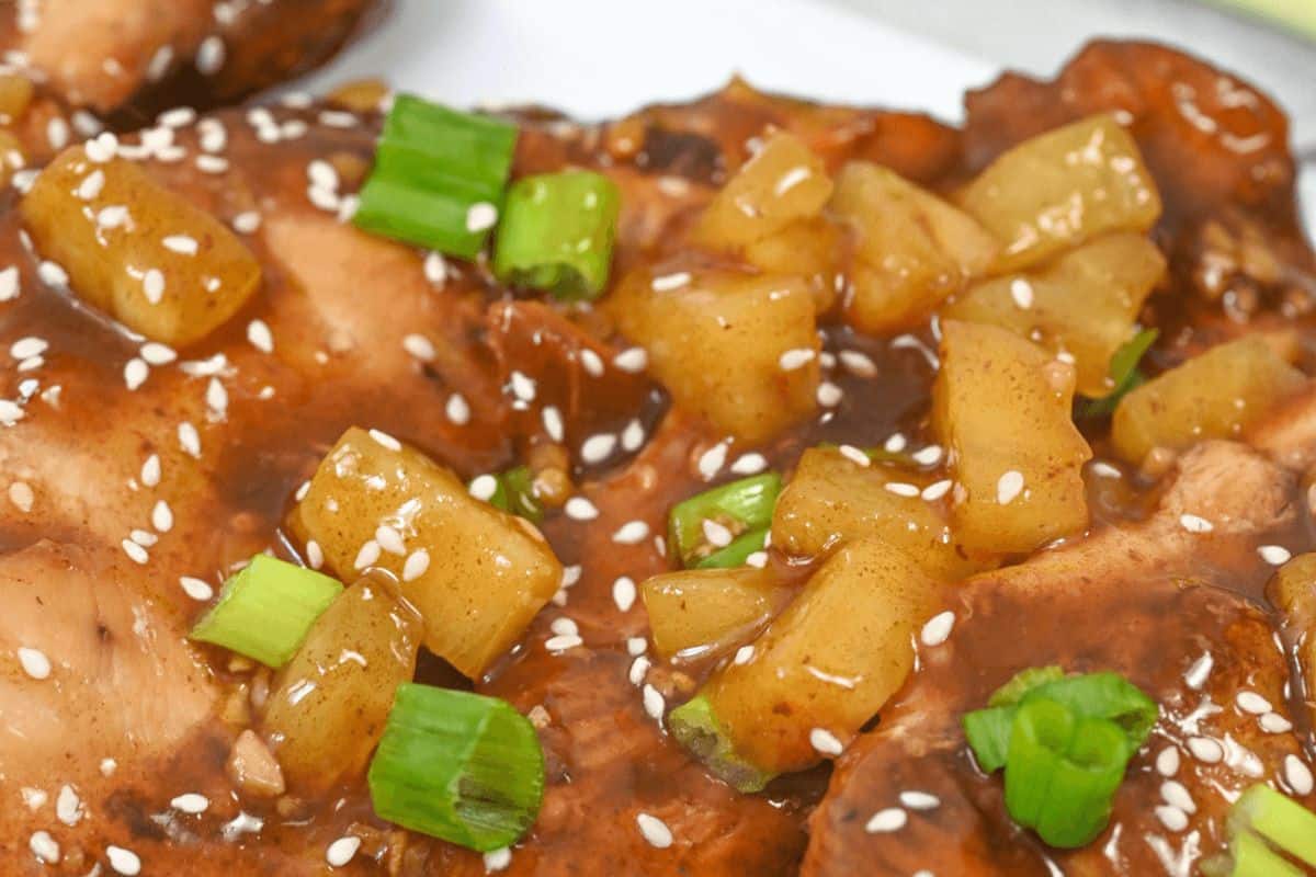 A very close up of chicken covered in a sauce that looks like the consistency of a sweet and sour sauce. The chicken has pineapple chunks and topped with sliced green onions and sesame seeds. 