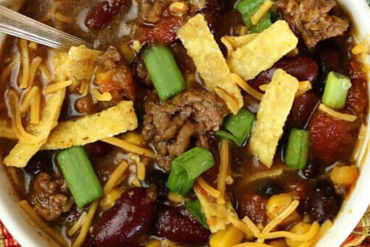A close up of a bowl of chili that is topped with grated cheese, tortilla strips and green onions. Tasty ground beef is visible and red beans. In the top left hand of the picture a silver utensil has been inserted into the chili.  