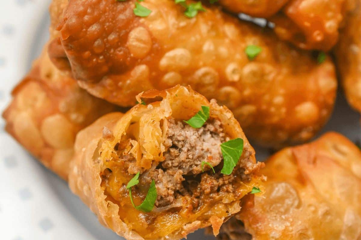 A very close up picture of egg rolls. The egg roll closest to the foreground of the picture has been cut so that you can see inside it. Ground beef, cheese and green herbs are visible in the closest egg roll. 