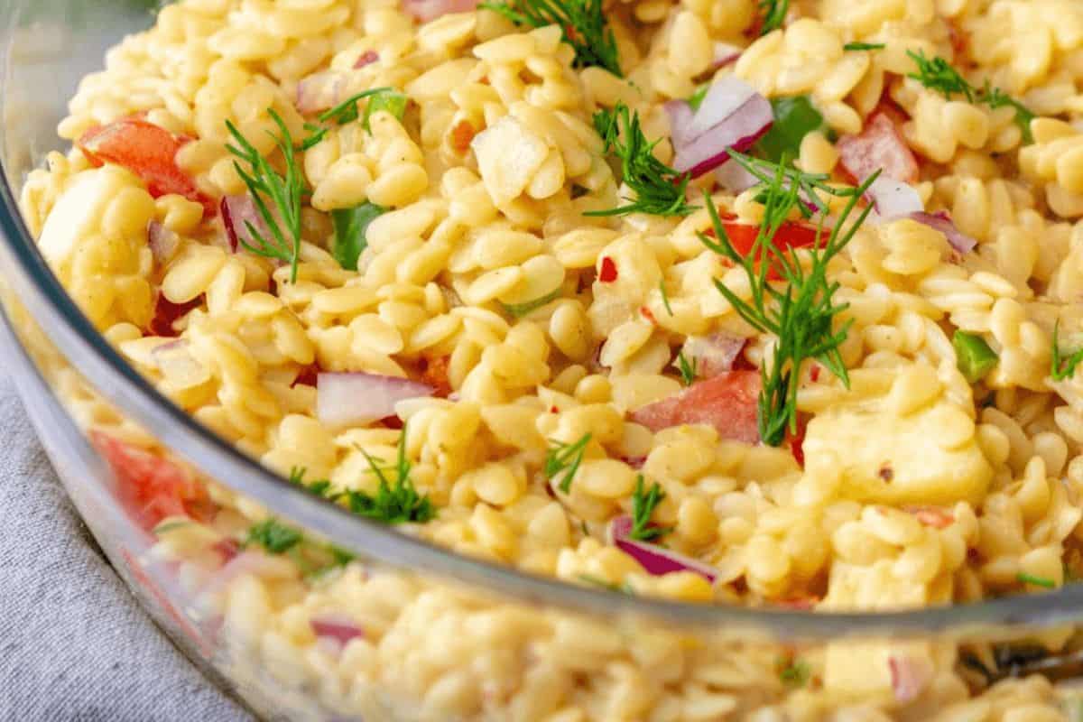 In a glass bowl, orzo is mixed with yellow corn, red bell pepper chunks, red onion slices and bright green sprigs of herbs. 