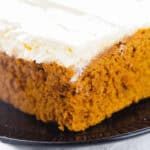 Close-up of a slice of moist pumpkin cake with a thick layer of cream cheese frosting on a black plate, evoking the comforting aromas reminiscent of pumpkin spice cake.