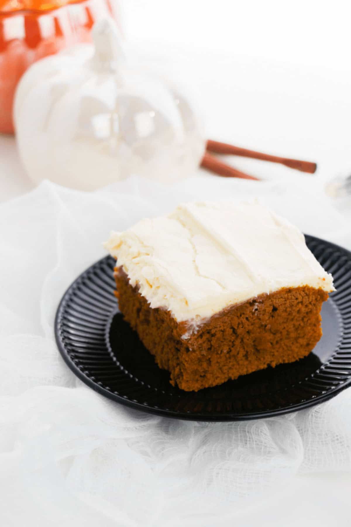 A slice of frosted pumpkin spice cake on a black plate with white pumpkin decor and cinnamon sticks in the background.