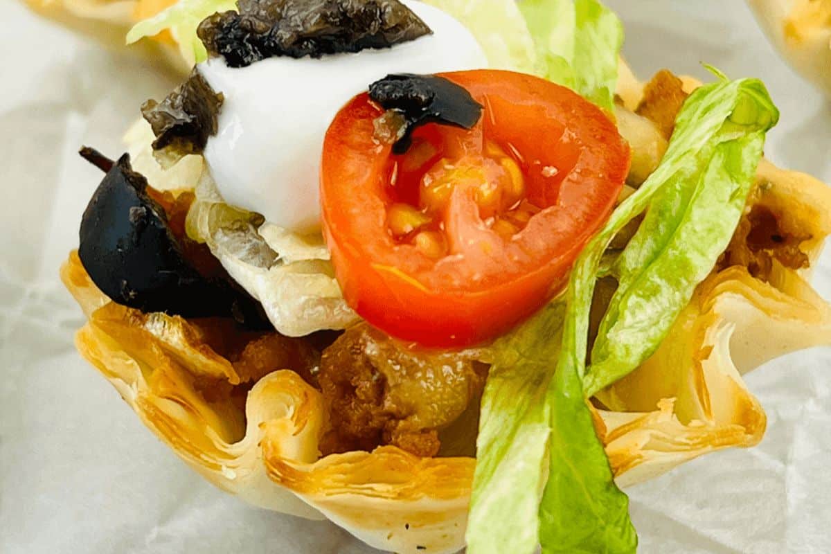 A very close up picture of a single Phyllo dough shell that is filled with ground beef, cheese, lettuce,  a cherry tomato, a black olive and topped with sour cream. 