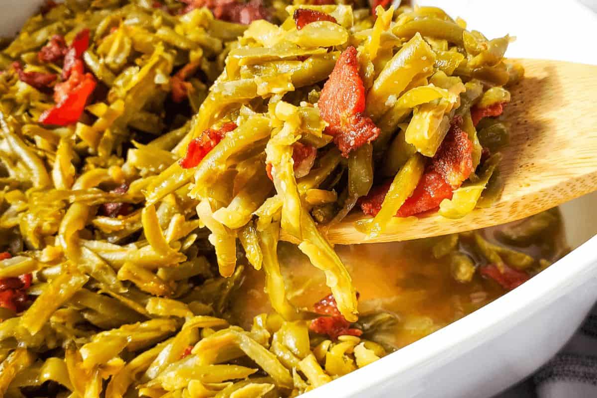 A white bowl of bacon green beans with a wooden spoon in them. 
