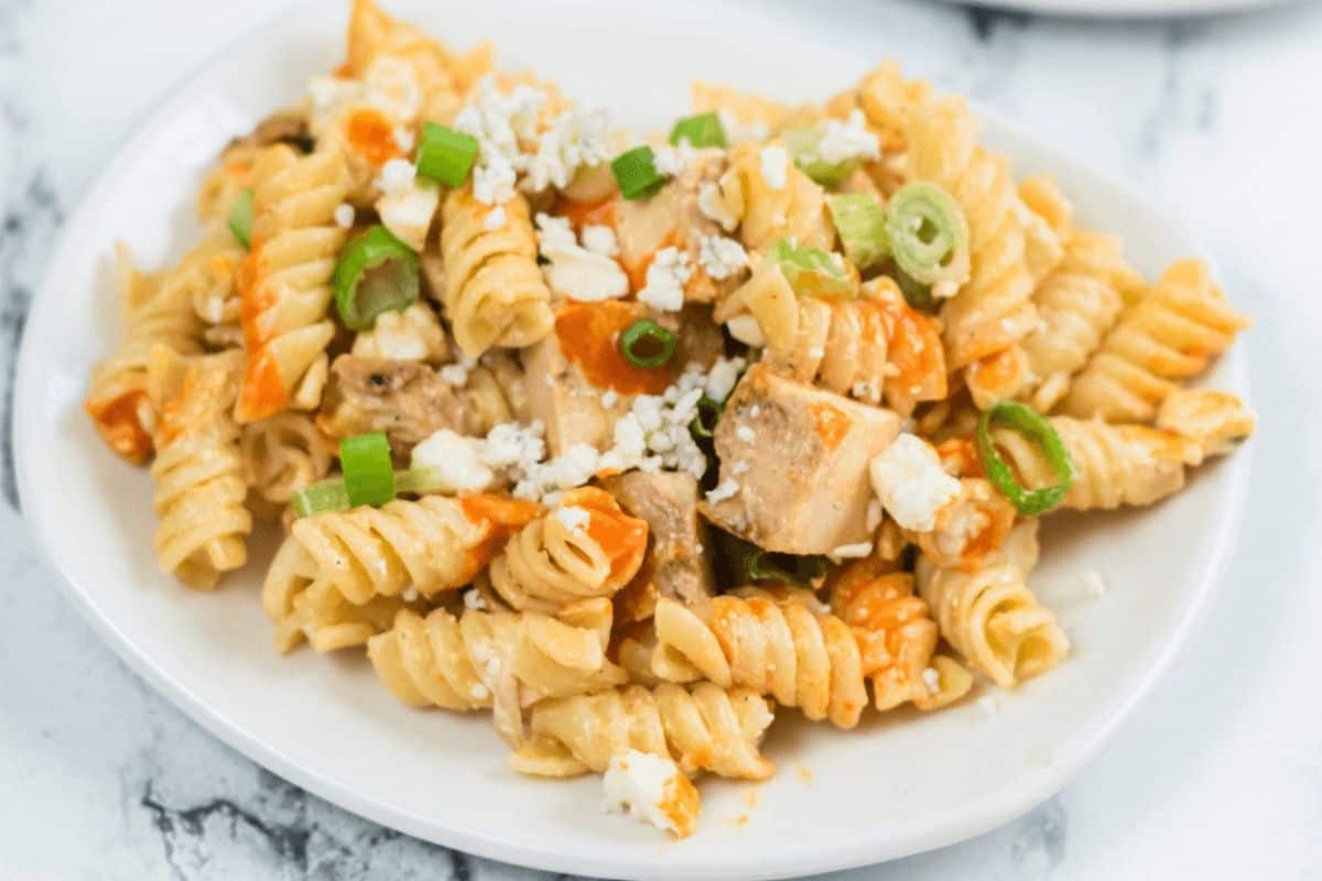 On a gray marbled countertop, sits a white plate that has cubes of chicken and rotini pasta on it. The chicken and pasta are coated with a light orange buffalo wing sauce. The dish is garnished with chopped up green onions and Blue cheese. 