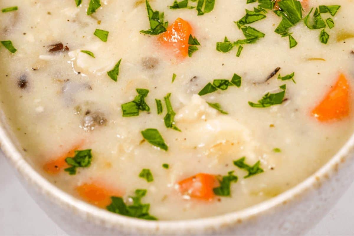 A close up of light yellow soup in a bowl. There are visible vegetables including carrots and parsley. 