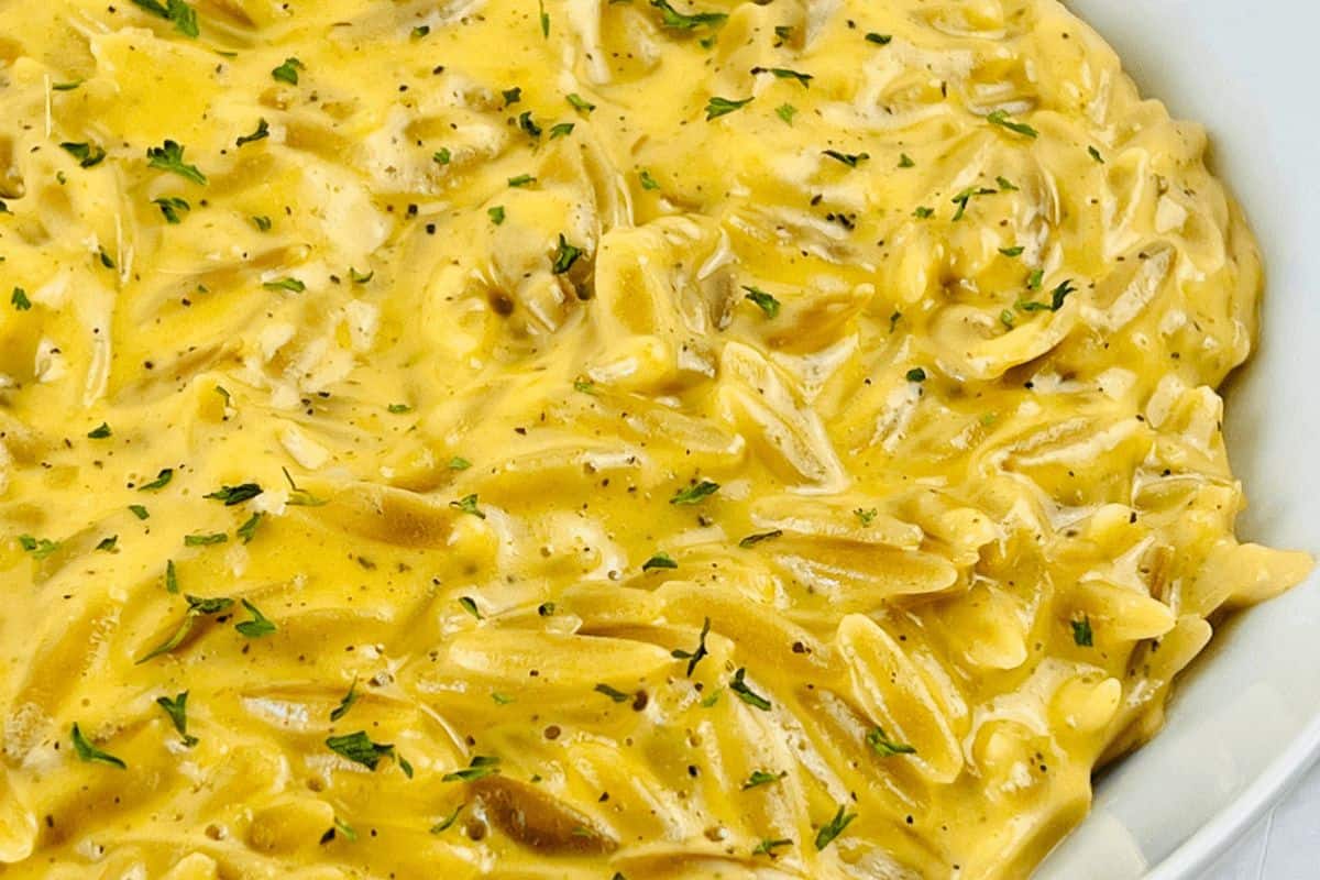 On a white background, there is a very close up picture of orzo in a yellow, cheesy sauce and topped with green herbs. 