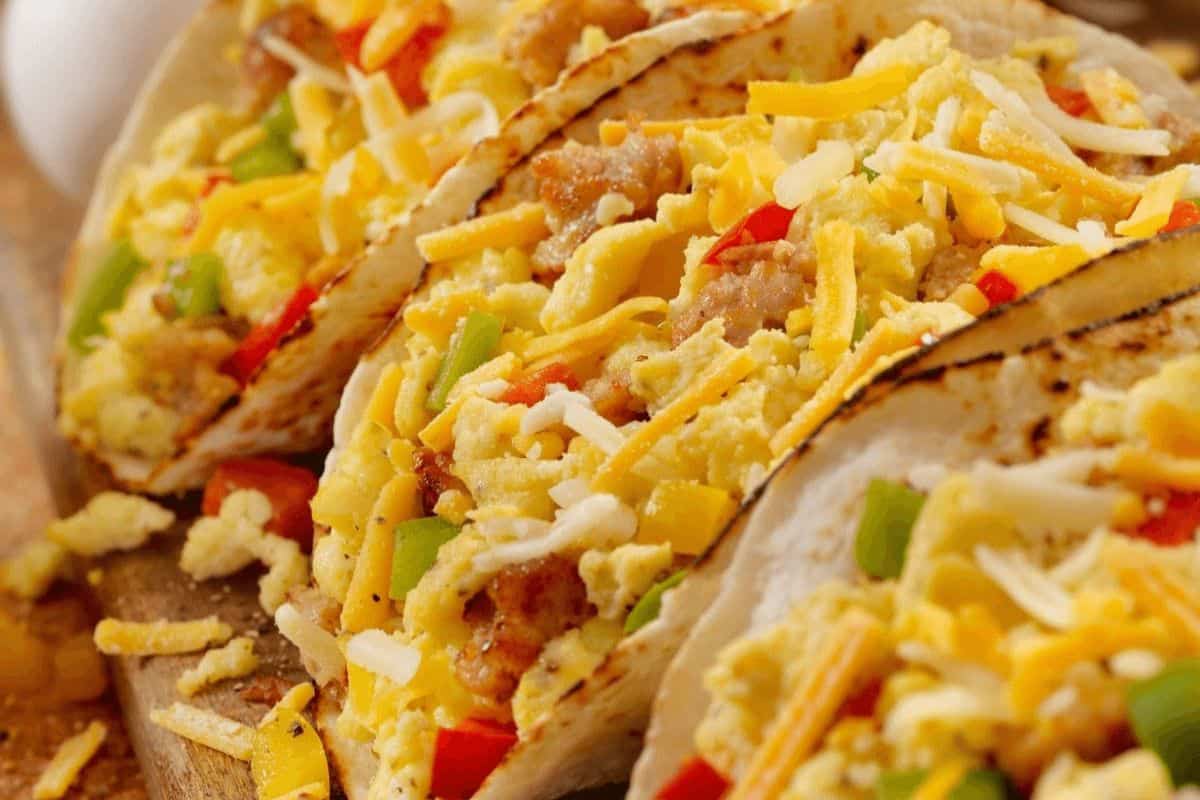 A close up of three tacos stacked horizontally with the middle taco in focus. The edges of the tacos are browned and are filled generously with eggs, breakfast sausage, cheese, diced tomatoes and cilantro. 