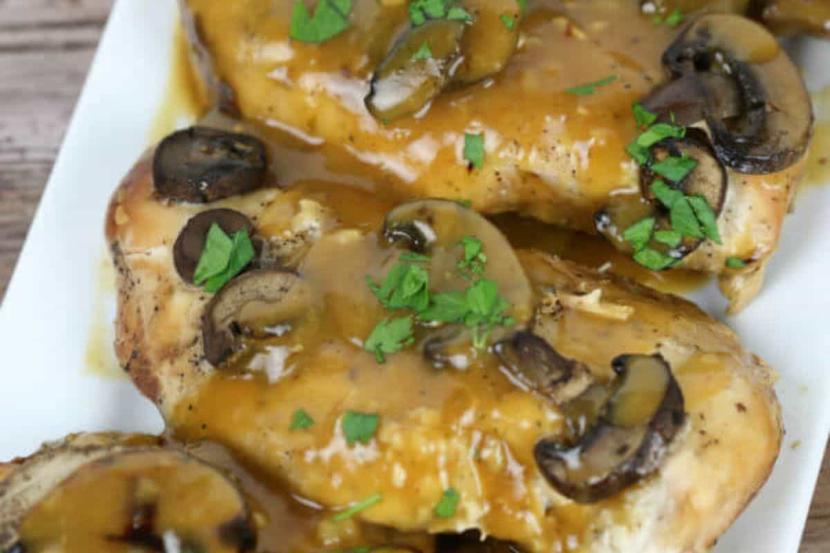 A close up of three chicken breasts covered in a honey colored sauce and topped with mushrooms and parsley. 
