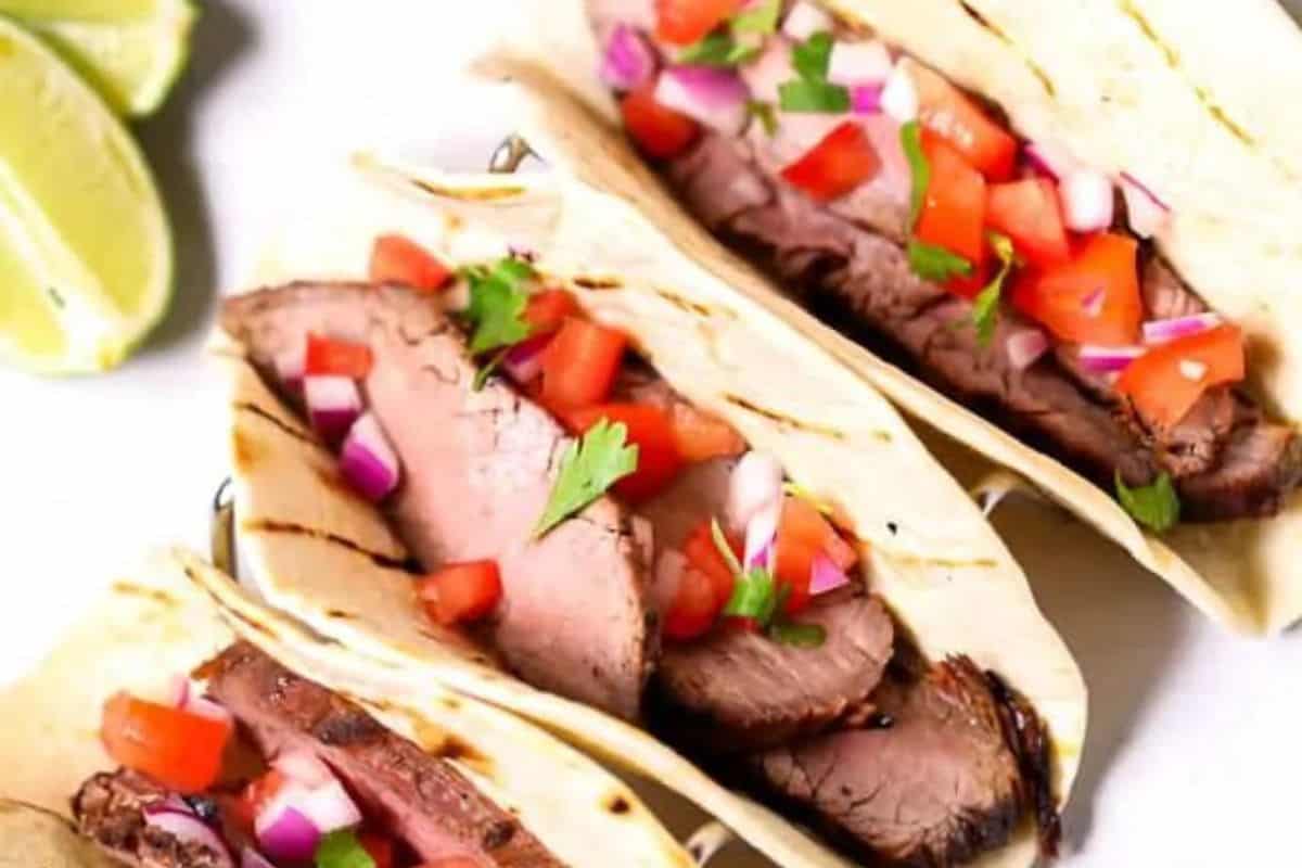On a white plate, three tacos stacked horizontally with wedges of lime decorating the plate in the top left corner. There are grill marks on the tacos and they are filled with strips of steak, diced tomatoes, purple onions and cilantro. 