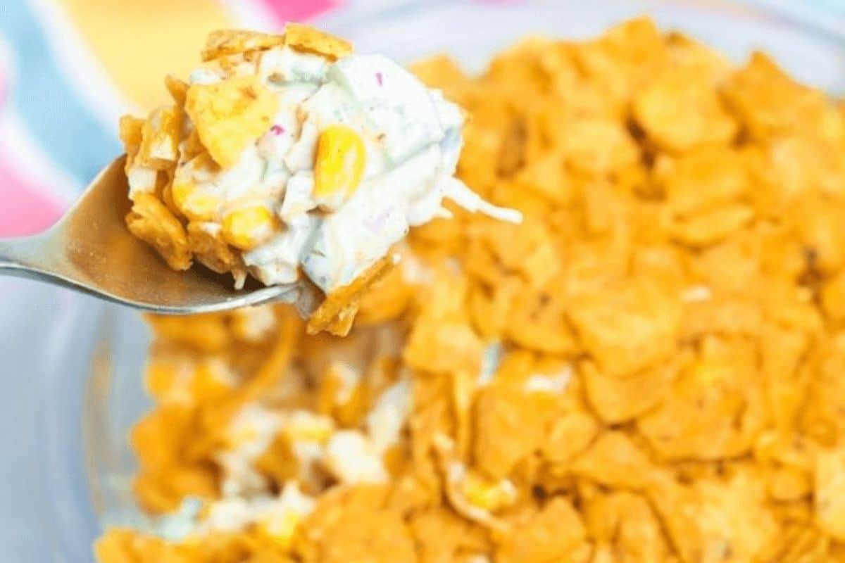 In the background there is a blurry dish filled with frito salad. In the foreground, there is a silver spoon with a spoonful of frito salad. The salad is creamy with corn, peppers, onions and topped with Fritos. 