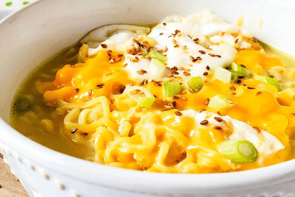 In a white bowl, ramen noodles are topped with cheese, an egg, toasted sesame seeds and green onions. 
