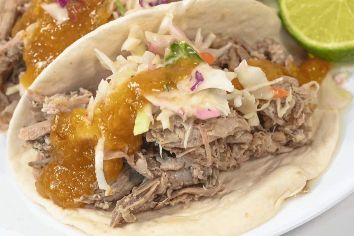 On a white plate, a filled taco with another one in the foreground and a wedge of lime on the right hand side of the plate. The taco is filled with pork and topped with coleslaw and peach barbecue sauce. 