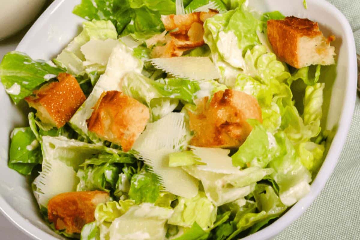 In a white bowl, a caesar salad fills the bowl. The lettuce is bright green and looks crispy, topped with homemade looking croutons and shaved cheese. 