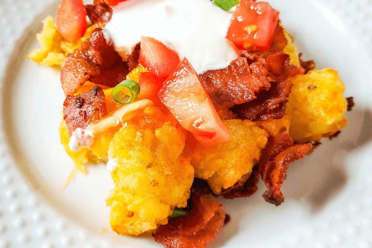 A very close up view of loaded tater tots on a white plate. The tater tots look crispy and are topped with sour cream, tomatoes, crispy bacon and slices of green onion. 