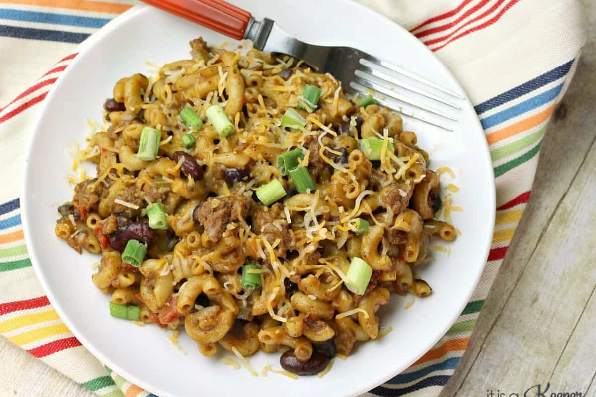 On a striped tea cloth a white plate is sitting with a mixture of mac and cheese with red kidney beans and tomatoes visible. The mac and cheese is topped with finely grated cheese and green onions. 