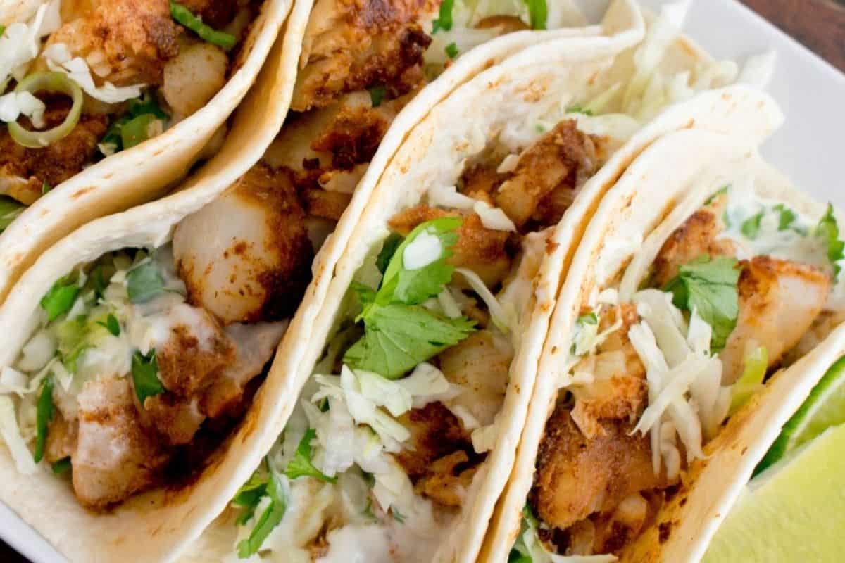 On a white plate, four tacos are tightly stacked horizontally with wedges of lime decorating the plate in the bottom right corner. The tacos are filled with grilled white fish, onion, cilantro and a white dressing. 