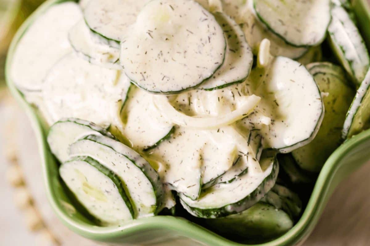 In a scalloped green bowl, there is a close up of cucumbers and onions tossed in a white dressing. The dressing looks creamy and has generous amounts of dill mixed in. 