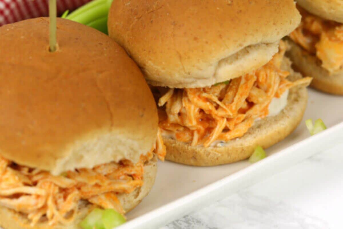On a white plate, there is a close up of two small burger buns filled with shredded chicken mixed in a red buffalo sauce. The bottom of the buns have blue cheese dressing. The foreground has little cubes of celery on the plate. 