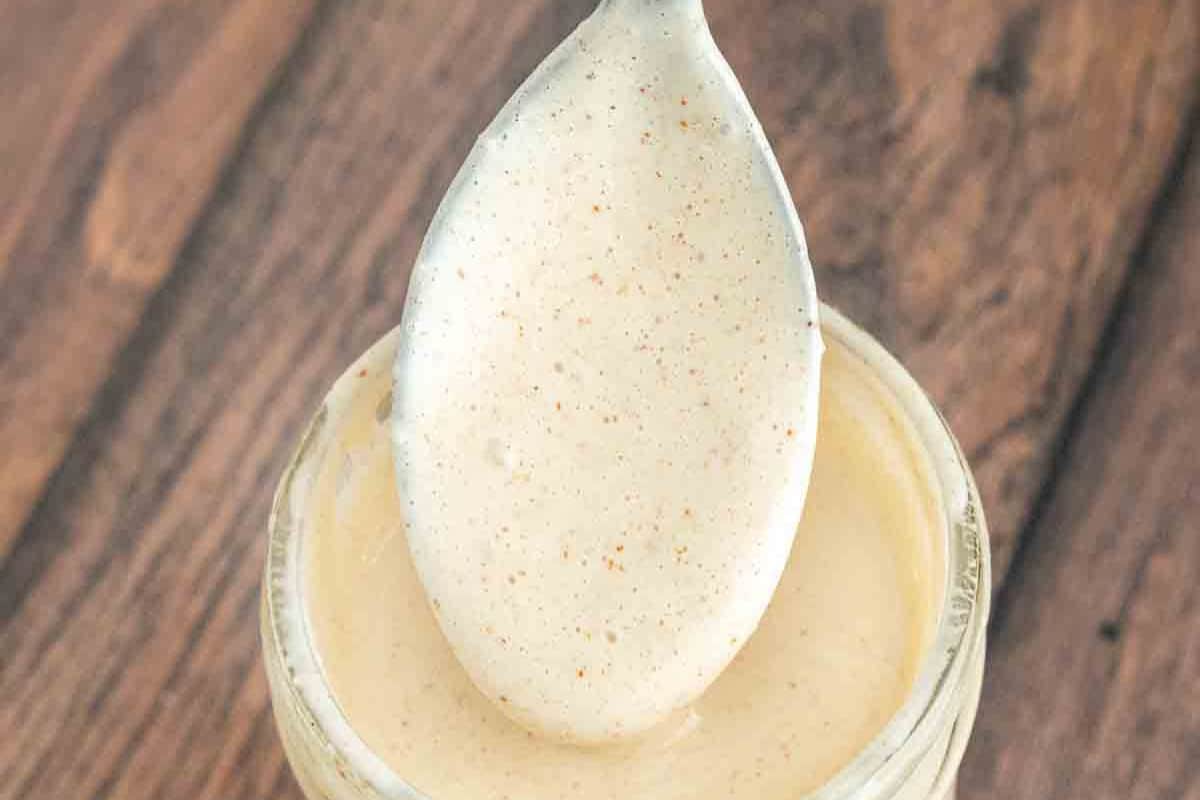 A spoon holding a thick, creamy, light-colored homemade BBQ sauce with visible specks, held over an open jar on a wooden surface.