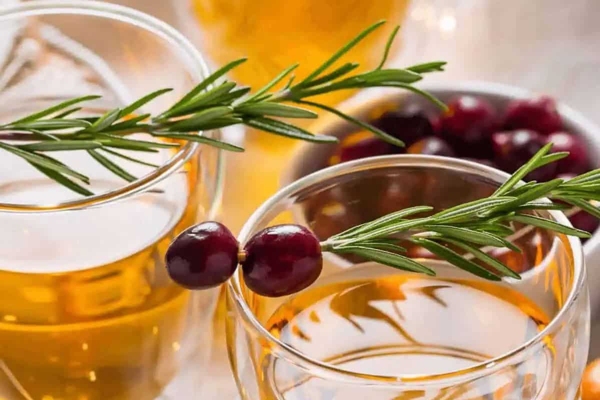 Glasses of golden apple boilo garnished with fresh rosemary sprigs and cranberries, with a bowl of cranberries in the background.