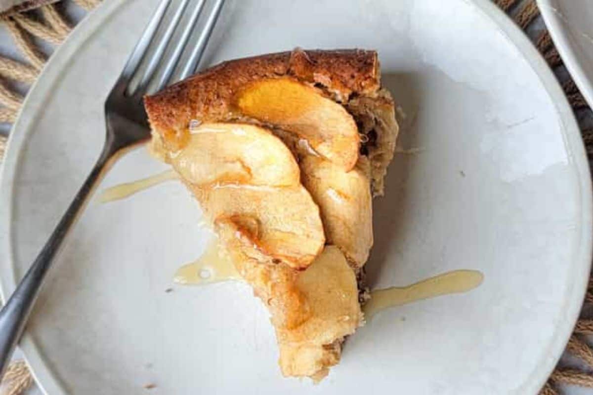 A slice of apple cake on a light gray plate with a shiny drizzle on top, next to a fork. Thin apple slices are visible on the golden crust.