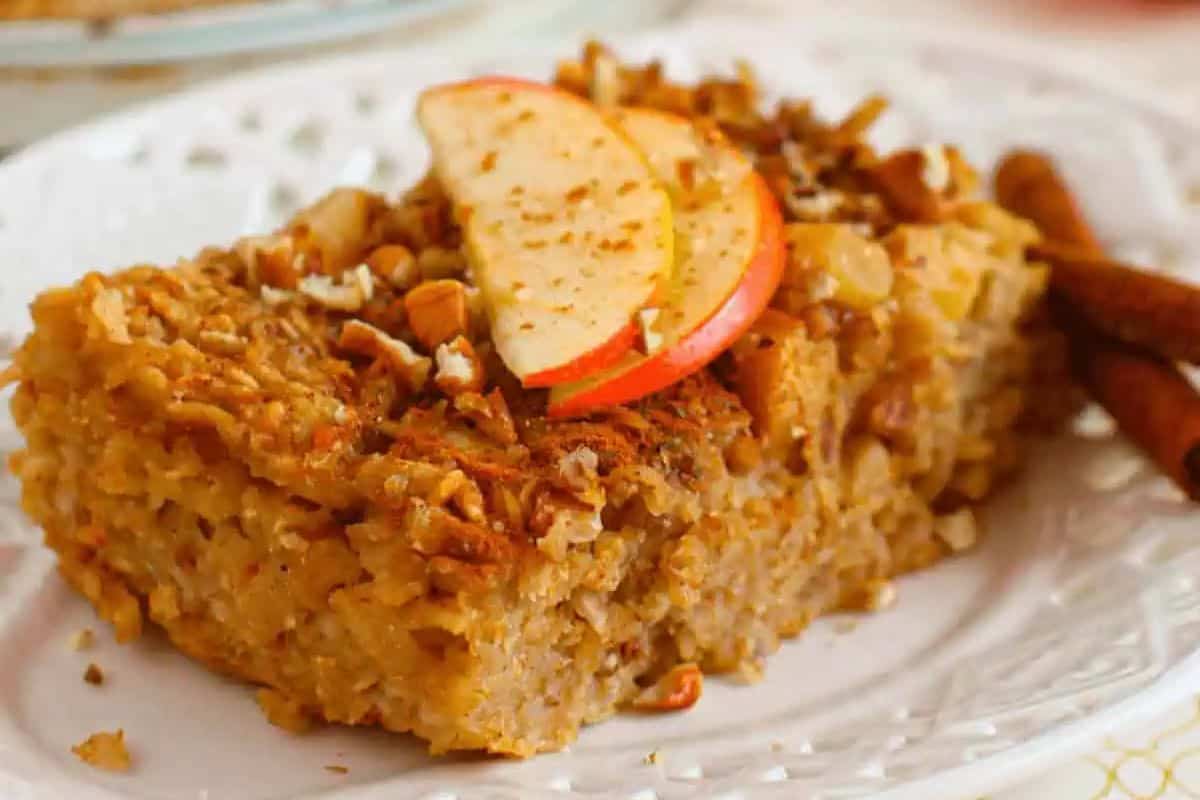 A slice of baked apple oatmeal topped with chopped nuts and fresh apple slices, served on a white plate with cinnamon sticks beside it.