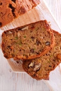 Two slices of banana zucchini bread with visible walnuts sit on a wooden cutting board, with part of a loaf in the background.
