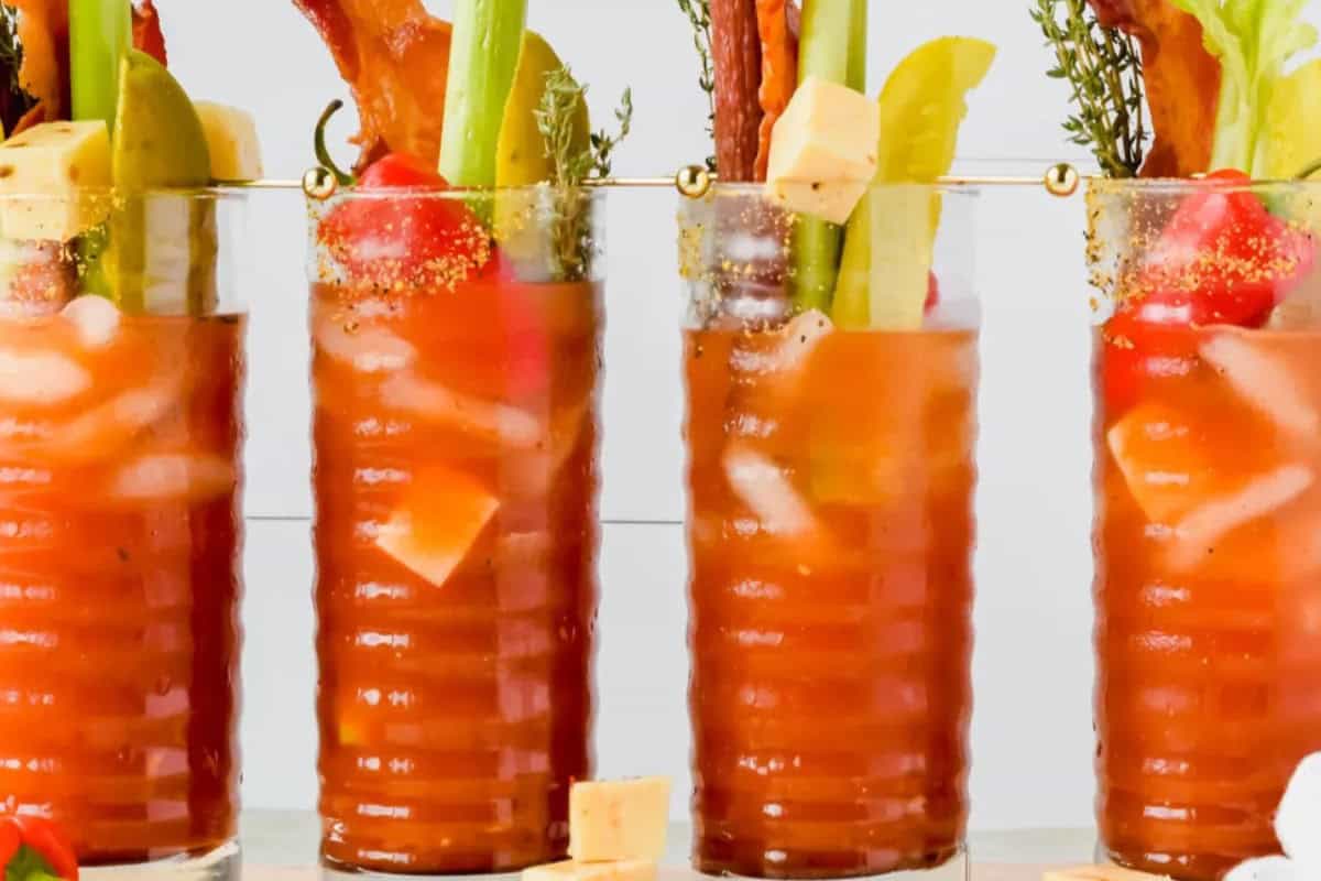 Three Bloody Mary cocktails—perfect for seasonal recipes—garnished with celery, bacon, cherry tomatoes, cheese cubes, and pickles, served in ribbed glasses with ice.