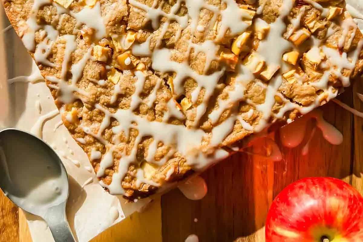 Close-up of apple blondies drizzled with white icing, resting on parchment paper with a spoon beside them and a fresh red apple below, all set on a wooden surface.
