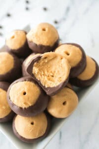 A bowl filled with buckeye candy, showcasing chocolate-dipped peanut butter balls, one with a bite taken, displayed on a white marble surface.